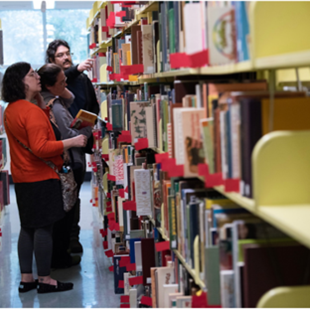 Three people stand in a brightly lit library aisle between yellow bookshelves with red book supports. One person in a red top browses the shelves, while the other two hold books and discuss a title, with one pointing at a book. Natural light streams in from a nearby window, creating a warm, collaborative atmosphere of shared learning.